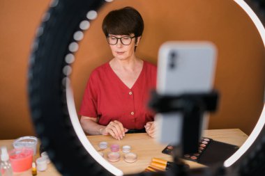 Middle-aged woman talking on cosmetics with makeup eye shadows and blush palette and brushes while recording her video. Mature female making video for her blog on cosmetics