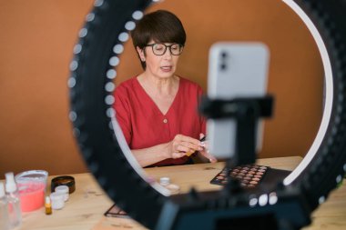Beauty and cosmetics e-commerce and blogging concept - professional mature female make up artist testing new cosmetics recording video review on cosmetic products at home