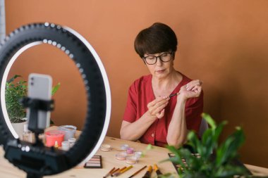 Middle-aged woman talking on cosmetics with makeup eye shadows and blush palette and brushes while recording her video. Mature female making video for her blog on cosmetics