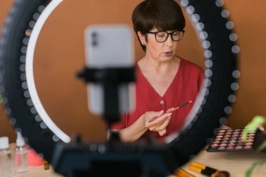 Beauty and cosmetics e-commerce and blogging concept - professional mature female make up artist testing new cosmetics recording video review on cosmetic products at home