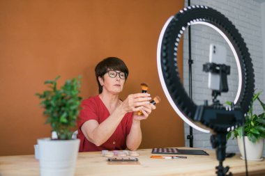 Middle-aged woman talking on cosmetics with makeup eye shadows and blush palette and brushes while recording her video. Mature female making video for her blog on cosmetics