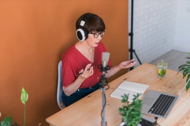 Mature woman making podcast recording for her online show. Attractive business woman using headphones front of microphone for a radio broadcast
