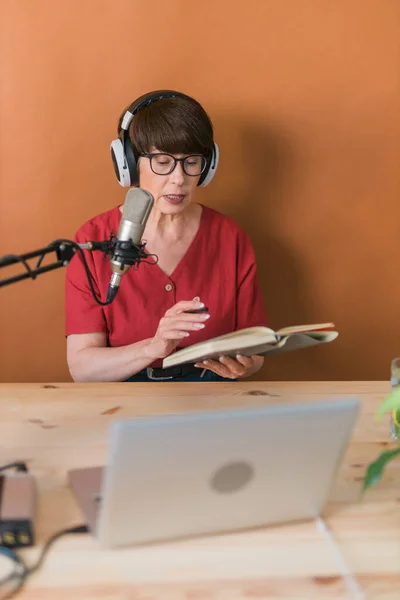 Mature woman making podcast recording for her online show. Attractive business woman using headphones front of microphone for a radio broadcast — Stock fotografie