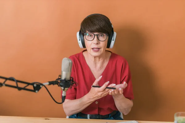 Mature woman making podcast recording for her online show. Attractive business woman using headphones front of microphone for a radio broadcast — Stock fotografie