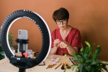 Middle-aged woman talking on cosmetics with makeup eye shadows and blush palette and brushes while recording her video. Mature female making video for her blog on cosmetics