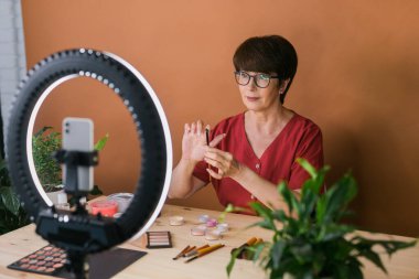 Beauty and cosmetics e-commerce and blogging concept - professional mature female make up artist testing new cosmetics recording video review on cosmetic products at home