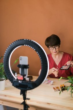 Middle-aged woman talking on cosmetics with makeup eye shadows and blush palette and brushes while recording her video. Mature female making video for her blog on cosmetics