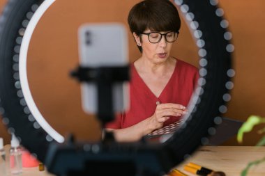Middle-aged woman talking on cosmetics with makeup eye shadows and blush palette and brushes while recording her video. Mature female making video for her blog on cosmetics