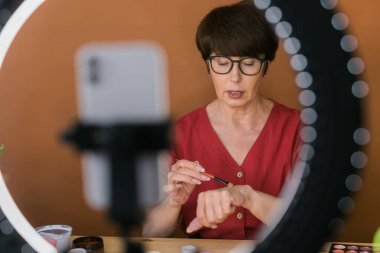 Beauty and cosmetics e-commerce and blogging concept - professional mature female make up artist testing new cosmetics recording video review on cosmetic products at home