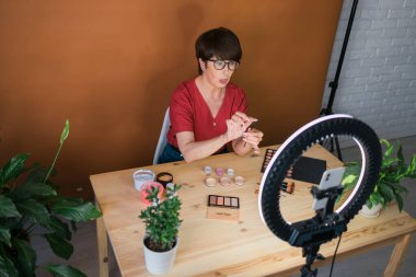 Beauty and cosmetics e-commerce and blogging concept - professional mature female make up artist testing new cosmetics recording video review on cosmetic products at home