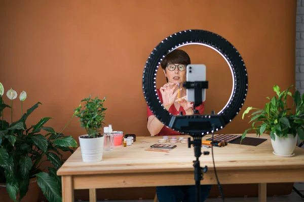 Middle-aged female beauty blogger or influencer with makeup product recording video in room at home. Using ring lamp and smartphone