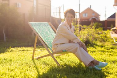 Young woman in pajama is resting in chair on a green lawn on sunny summer day - village and country life