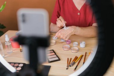 Middle-aged woman talking on cosmetics with makeup eye shadows and blush palette and brushes while recording her video. Mature female making video for her blog on cosmetics