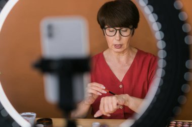 Middle-aged woman talking on cosmetics with makeup eye shadows and blush palette and brushes while recording her video. Mature female making video for her blog on cosmetics
