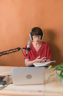 Mature woman making podcast recording for her online show. Attractive business woman using headphones front of microphone for a radio broadcast