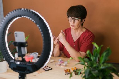 Middle-aged woman talking on cosmetics with makeup eye shadows and blush palette and brushes while recording her video. Mature female making video for her blog on cosmetics