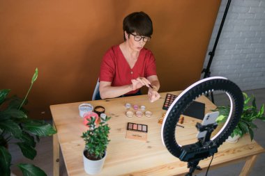 Beauty and cosmetics e-commerce and blogging concept - professional mature female make up artist testing new cosmetics recording video review on cosmetic products at home