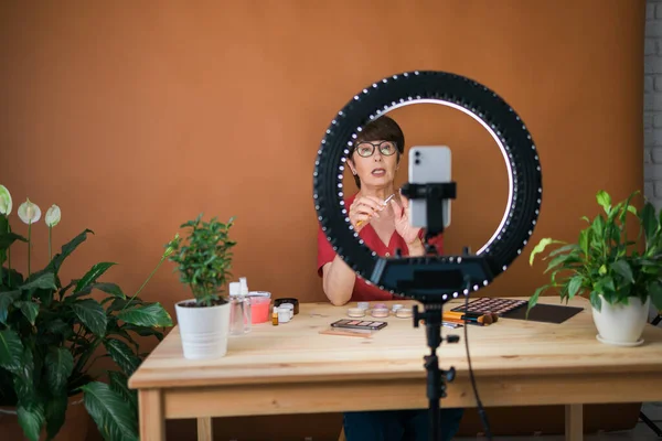 Middle-aged woman talking on cosmetics with makeup eye shadows and blush palette and brushes while recording her video. Mature female making video for her blog on cosmetics — Stock fotografie