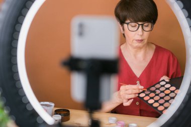 Beauty and cosmetics e-commerce and blogging concept - professional mature female make up artist testing new cosmetics recording video review on cosmetic products at home