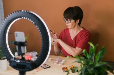 Beauty and cosmetics e-commerce and blogging concept - professional mature female make up artist testing new cosmetics recording video review on cosmetic products at home
