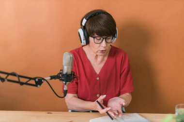 Mature woman making podcast recording for her online show. Attractive business woman using headphones front of microphone for a radio broadcast