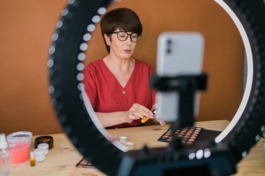 Beauty and cosmetics e-commerce and blogging concept - professional mature female make up artist testing new cosmetics recording video review on cosmetic products at home