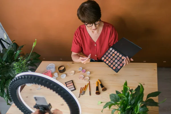 Beauty and cosmetics e-commerce and blogging concept - professional mature female make up artist testing new cosmetics recording video review on cosmetic products at home