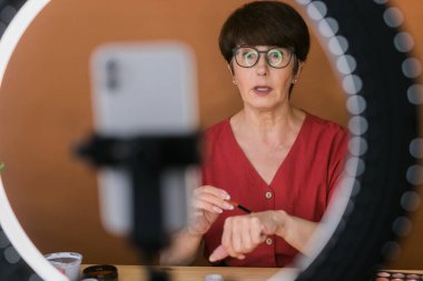 Middle-aged woman talking on cosmetics with makeup eye shadows and blush palette and brushes while recording her video. Mature female making video for her blog on cosmetics