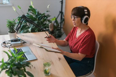 Mature woman making podcast recording for her online show. Attractive business woman using headphones front of microphone for a radio broadcast