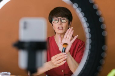 Middle-aged woman talking on cosmetics with makeup eye shadows and blush palette and brushes while recording her video. Mature female making video for her blog on cosmetics