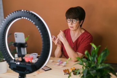 Beauty and cosmetics e-commerce and blogging concept - professional mature female make up artist testing new cosmetics recording video review on cosmetic products at home