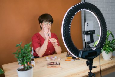 Beauty and cosmetics e-commerce and blogging concept - professional mature female make up artist testing new cosmetics recording video review on cosmetic products at home