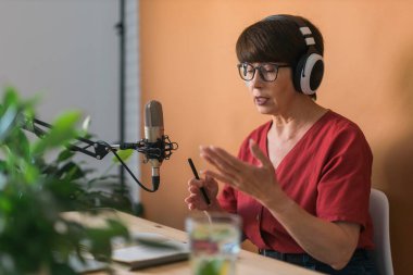 Mature woman making podcast recording for her online show. Attractive business woman using headphones front of microphone for a radio broadcast