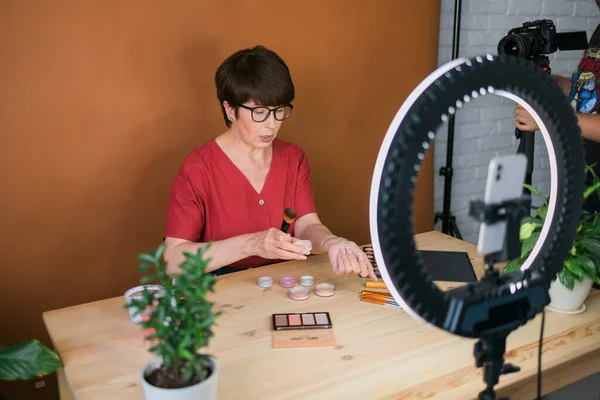 Middle-aged female beauty blogger or influencer with makeup product recording video in room at home. Using ring lamp and smartphone
