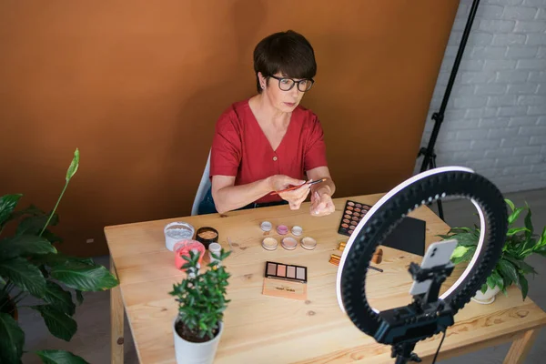 Beauty and cosmetics e-commerce and blogging concept - professional mature female make up artist testing new cosmetics recording video review on cosmetic products at home