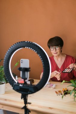 Middle-aged woman talking on cosmetics with makeup eye shadows and blush palette and brushes while recording her video. Mature female making video for her blog on cosmetics