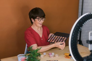 Beauty and cosmetics e-commerce and blogging concept - professional mature female make up artist testing new cosmetics recording video review on cosmetic products at home