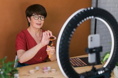 Middle-aged female beauty blogger or influencer with makeup product recording video in room at home. Using ring lamp and smartphone