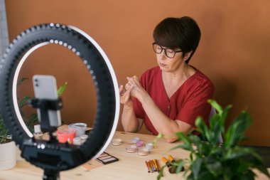 Beauty and cosmetics e-commerce and blogging concept - professional mature female make up artist testing new cosmetics recording video review on cosmetic products at home