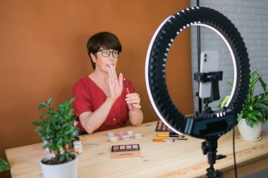 Beauty and cosmetics e-commerce and blogging concept - professional mature female make up artist testing new cosmetics recording video review on cosmetic products at home