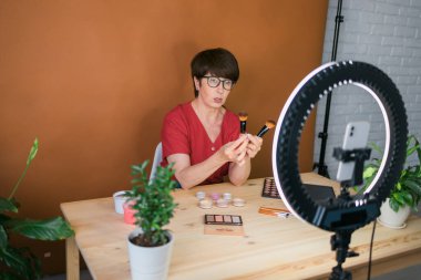 Middle-aged female beauty blogger or influencer with makeup product recording video in room at home. Using ring lamp and smartphone