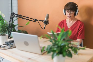 Mature woman making podcast recording for her online show. Attractive business woman using headphones front of microphone for a radio broadcast