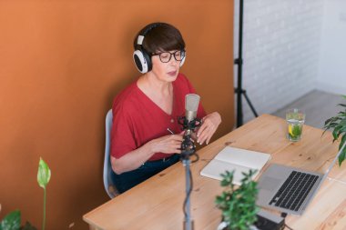 Mature woman making podcast recording for her online show. Attractive business woman using headphones front of microphone for a radio broadcast
