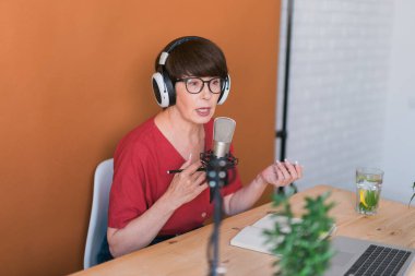 Mature woman making podcast recording for her online show. Attractive business woman using headphones front of microphone for a radio broadcast
