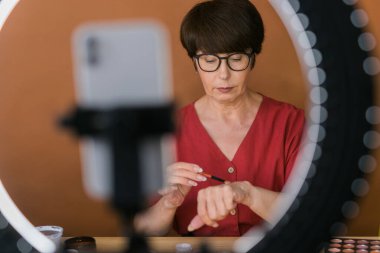 Beauty and cosmetics e-commerce and blogging concept - professional mature female make up artist testing new cosmetics recording video review on cosmetic products at home