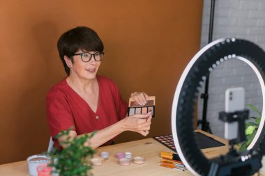 Middle-aged woman talking on cosmetics with makeup eye shadows and blush palette and brushes while recording her video. Mature female making video for her blog on cosmetics