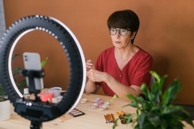 Middle-aged woman talking on cosmetics with makeup eye shadows and blush palette and brushes while recording her video. Mature female making video for her blog on cosmetics