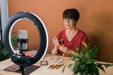 Beauty and cosmetics e-commerce and blogging concept - professional mature female make up artist testing new cosmetics recording video review on cosmetic products at home