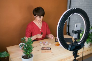 Middle-aged woman talking on cosmetics with makeup eye shadows and blush palette and brushes while recording her video. Mature female making video for her blog on cosmetics