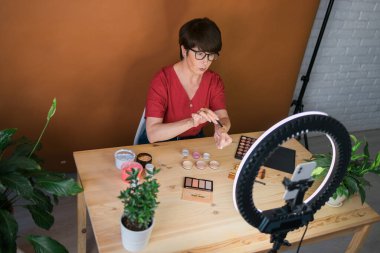 Beauty and cosmetics e-commerce and blogging concept - professional mature female make up artist testing new cosmetics recording video review on cosmetic products at home